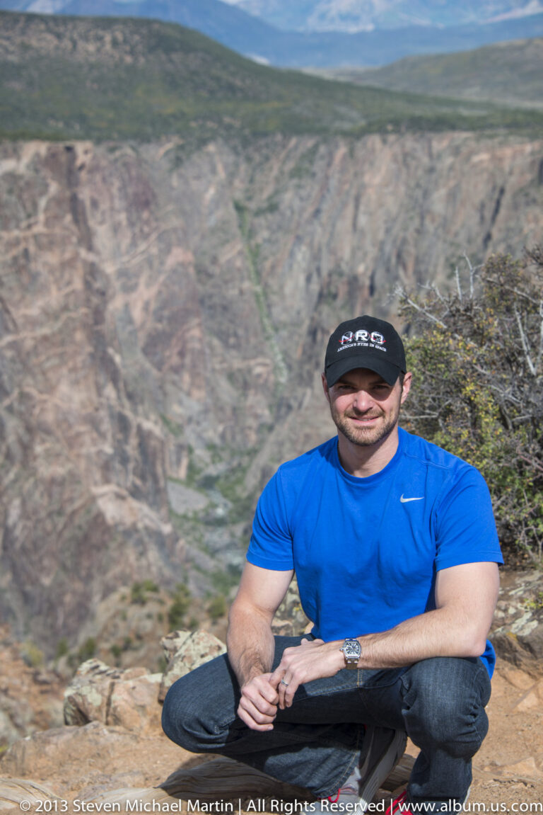 A man in a blue shirt and black cap sits on a rocky ledge with the canyon descending behind him.