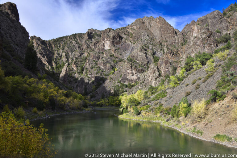 Green river water bends below steep canyon walls, with autumn foliage tracing the narrow shoreline.