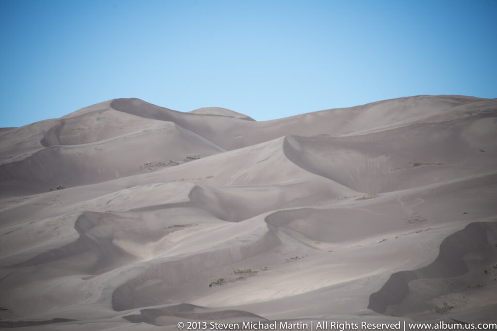 Pale sand ridges overlap in smooth curves beneath a clear blue sky.