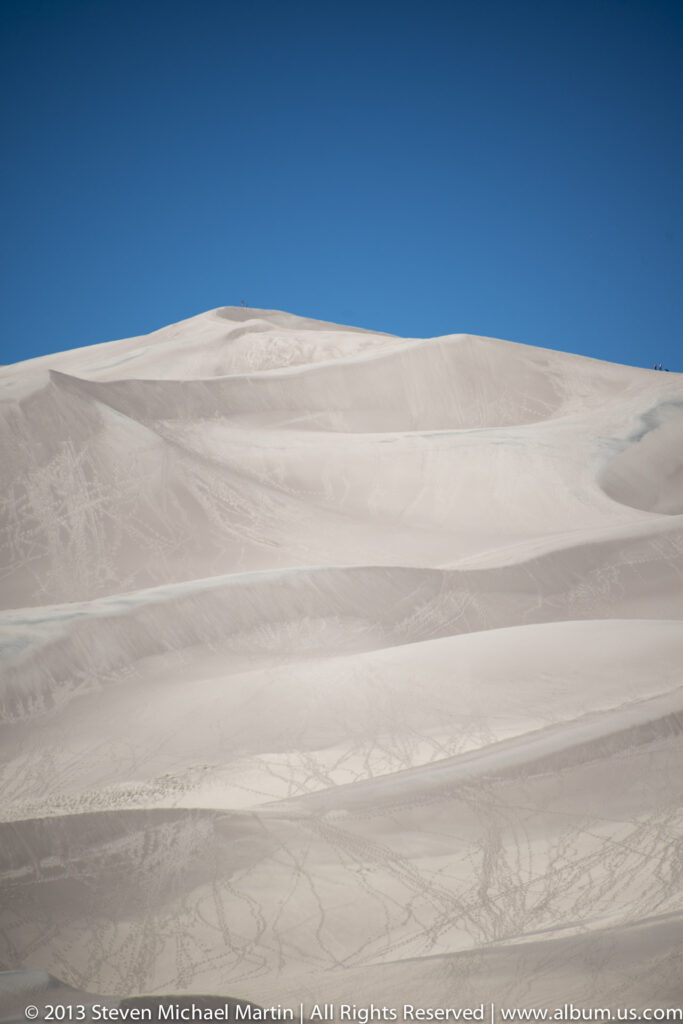 Pale dune slopes and shallow ridges sweep toward a low crest beneath blue sky.