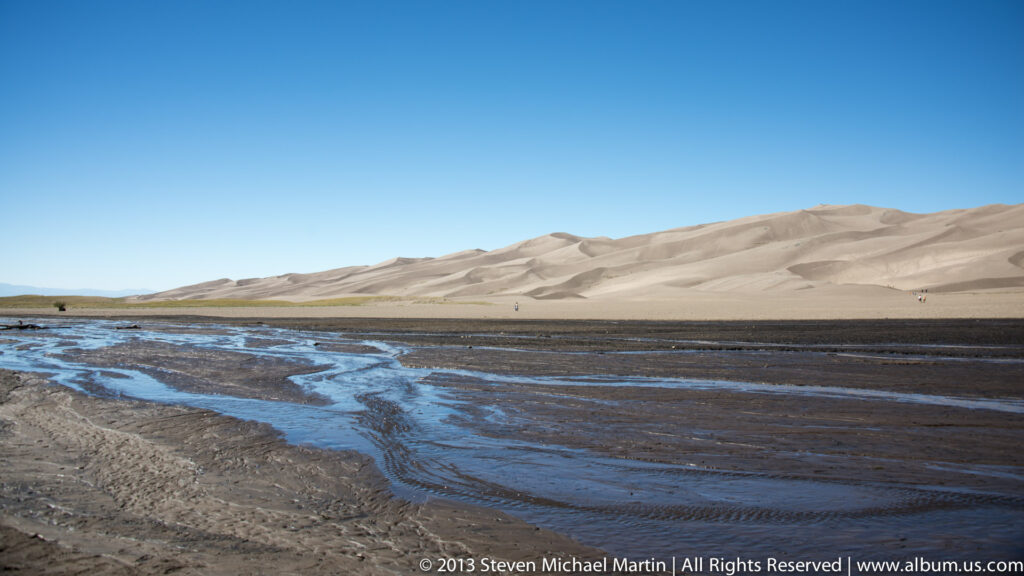 Shallow blue channels cross dark wet sand below a pale dune ridge with tiny visitors in the distance.