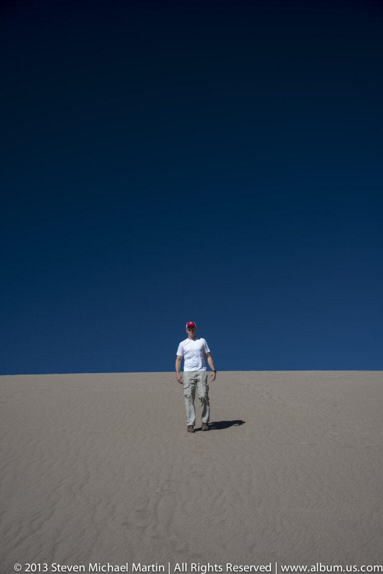 A small visitor walks away across a broad dune, casting a long shadow beneath a deep blue sky.