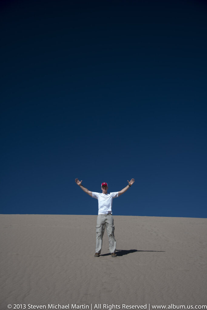 A man stands on a bare dune with both arms outstretched and a long shadow on the sand.