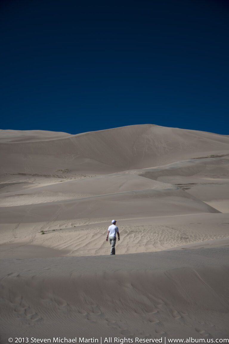 A small visitor walks down a broad smooth dune beneath a deep blue sky.
