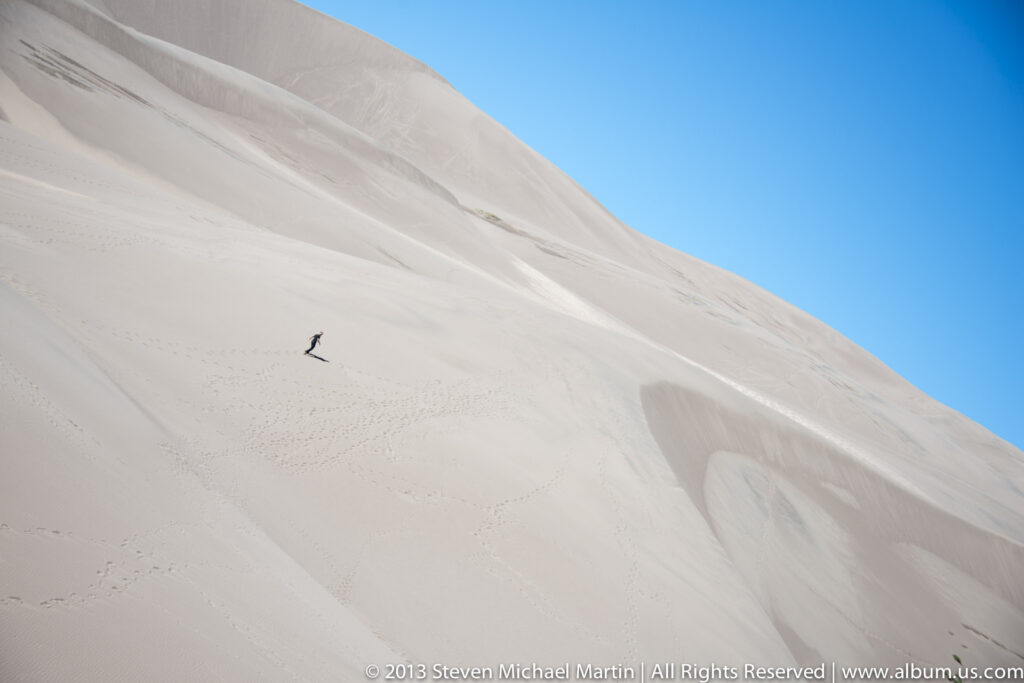 A small visitor crosses a steep pale dune marked by faint footprints beneath a blue sky.