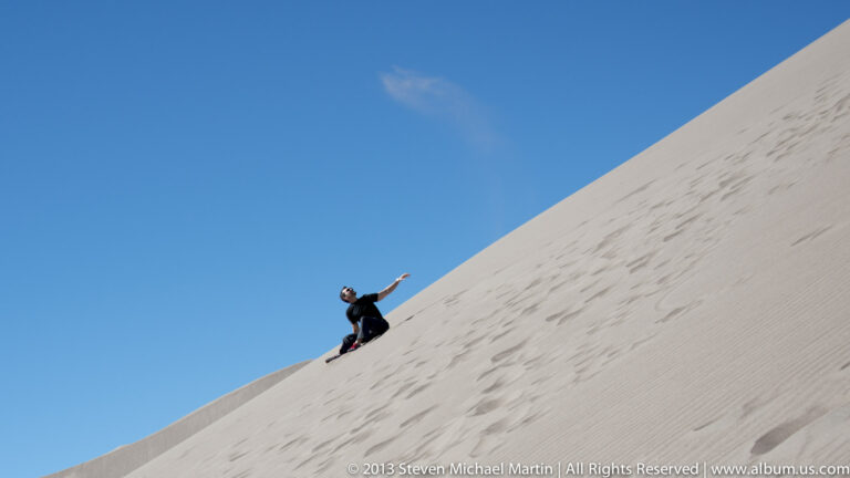 A person in dark clothing sits on a steep dune with one arm raised against a blue sky.