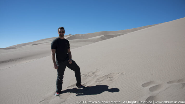 A man in dark clothing poses on a dune with one foot raised and footprints leading toward him.