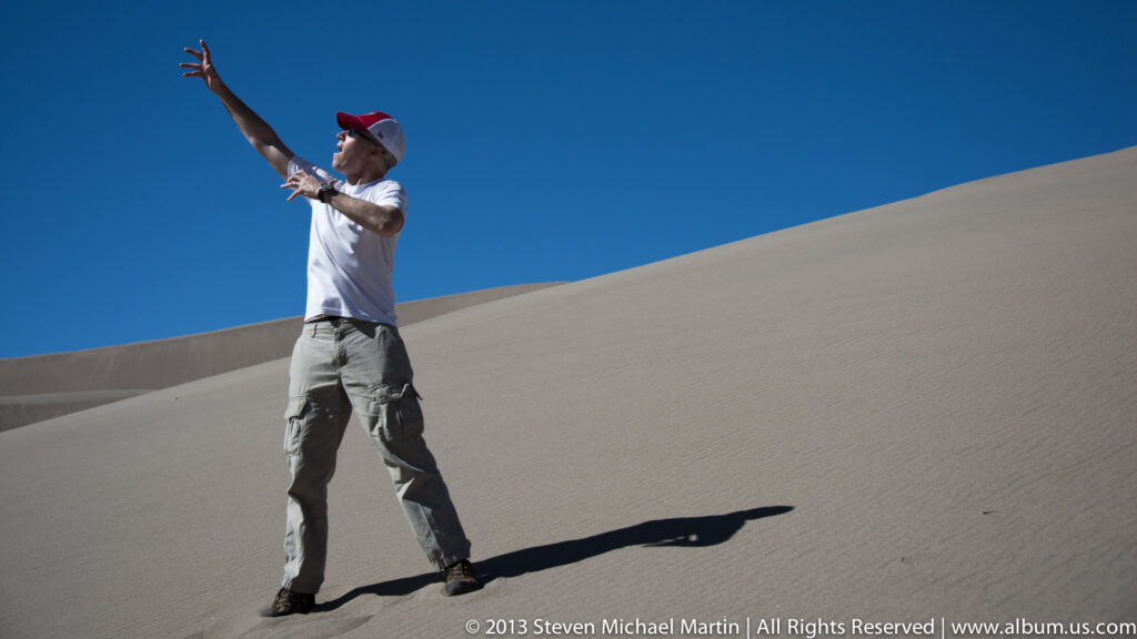 A man in a white shirt and red cap extends an arm while standing on a steep dune beneath blue sky.