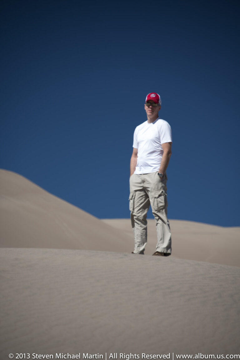 A man in a white shirt and red cap stands on a dune beneath a broad blue sky.