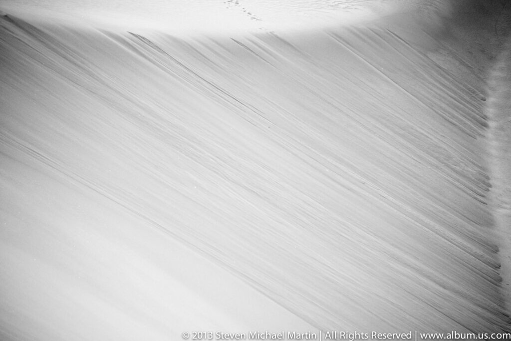Parallel ripples cross pale sand in alternating bands of light and shadow.