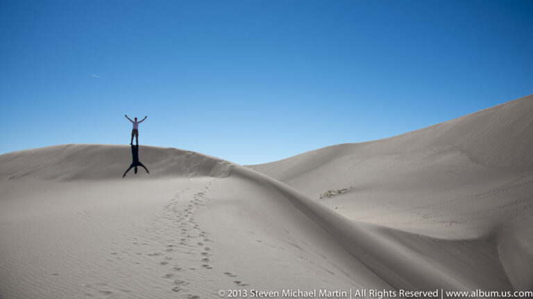 A visitor raises both arms at a dune crest reached by a trail of footprints.