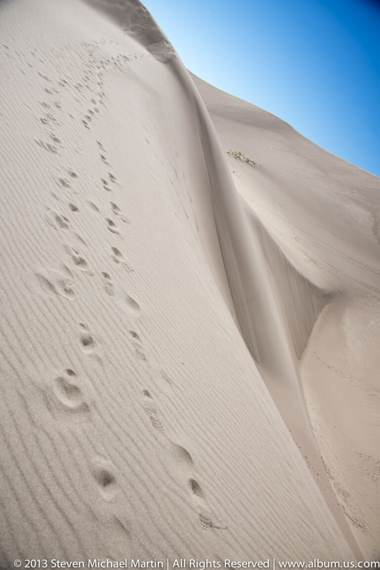 Footprints climb a steep pale dune toward a sharp crest beneath a narrow blue sky.