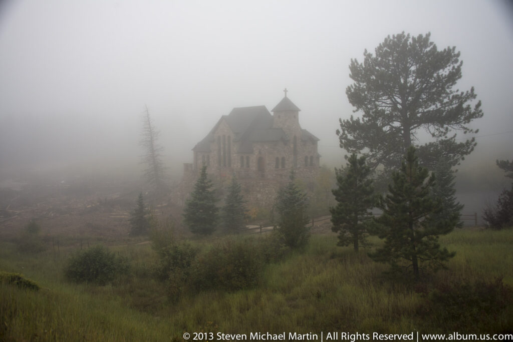 A stone chapel with a cross-topped tower emerges from fog among pine trees above a damp meadow.
