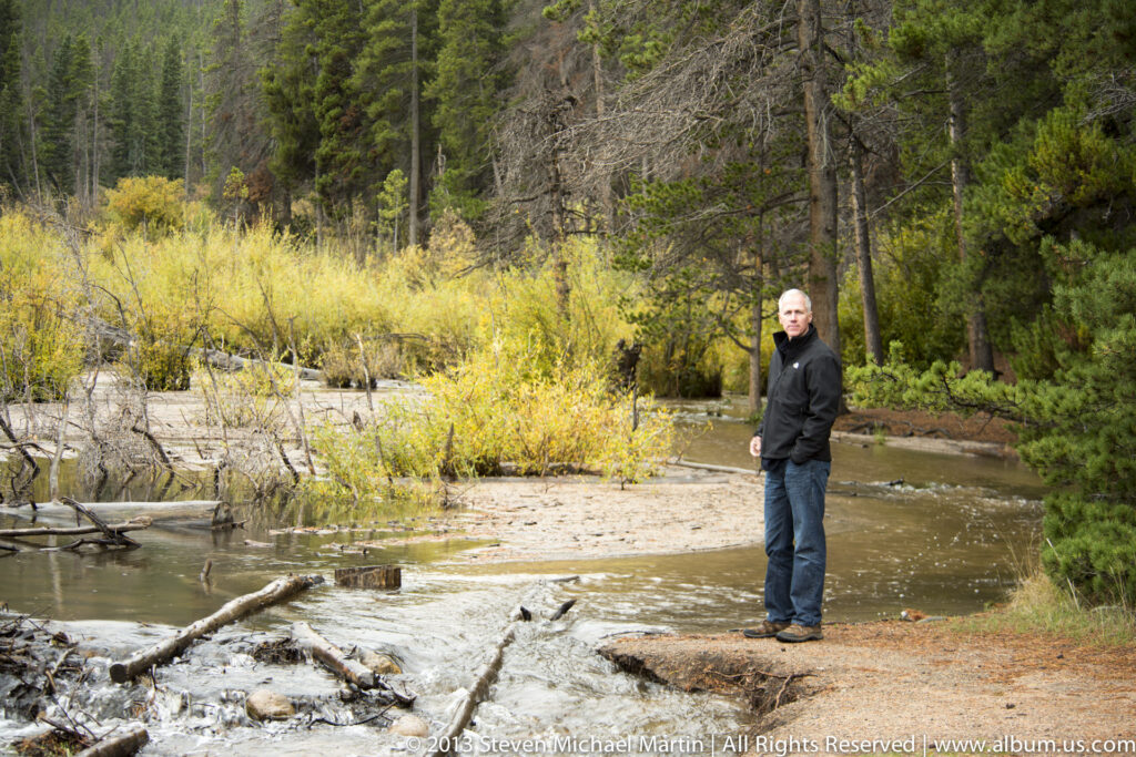 A man stands beside a shallow rocky stream bordered by yellow shrubs and evergreen trees.