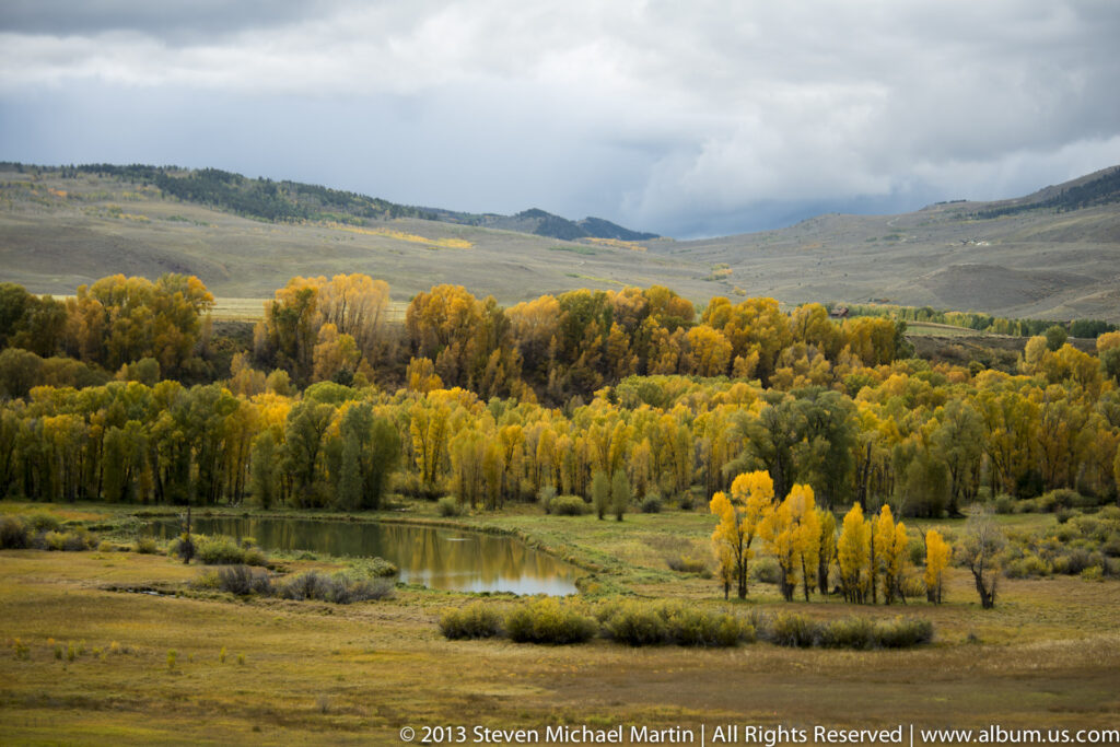 A reflective pond and yellow aspen grove sit below rolling grassy hills under a cloudy sky.
