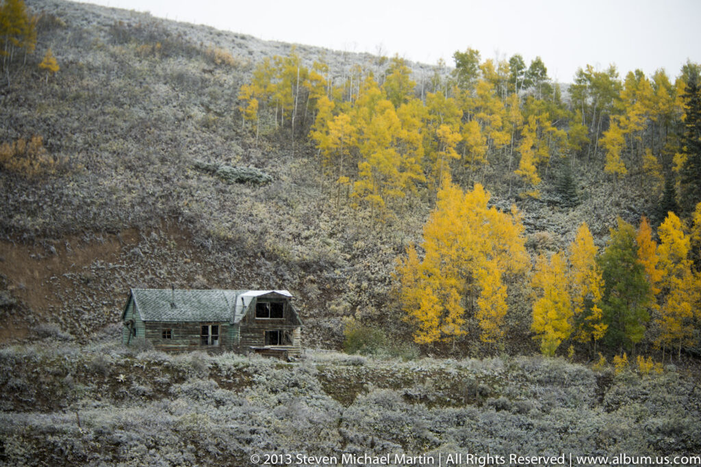 A deteriorating wooden cabin sits below yellow aspens on a steep, frost-dusted hillside.