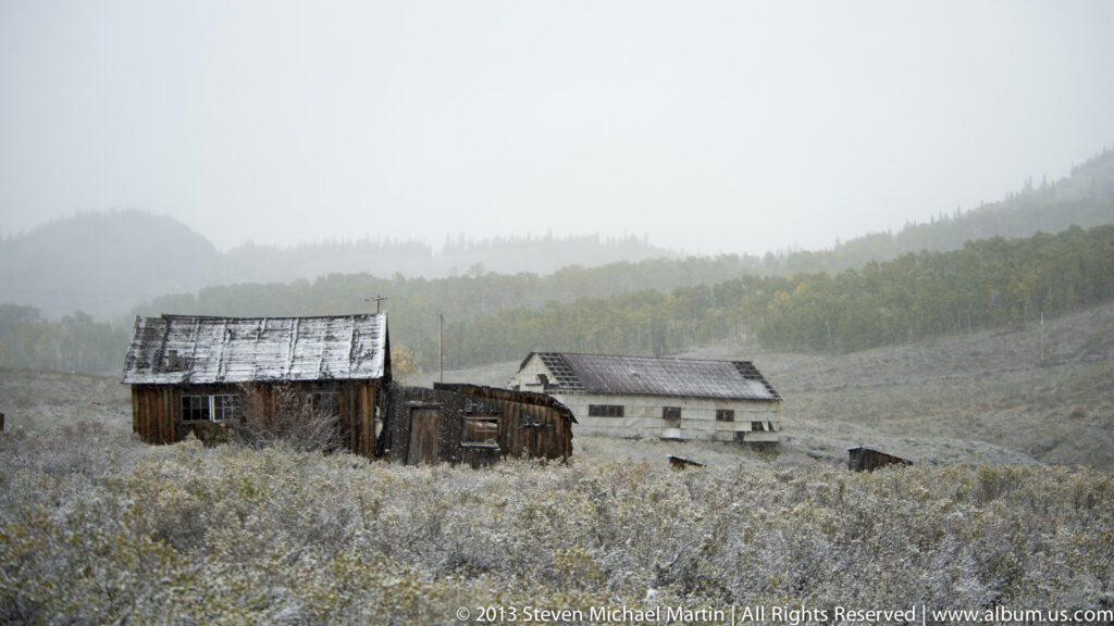 Two weathered cabins stand in frost-covered grass, with a fog-softened forested hillside behind them.