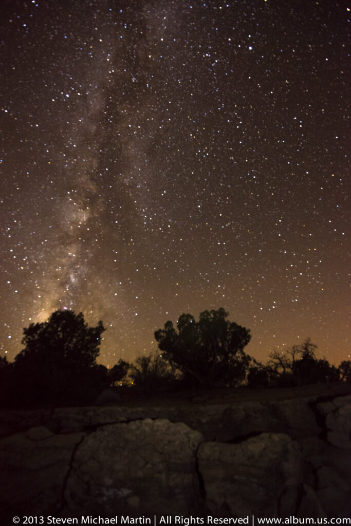 Milky Way and stars glow above silhouetted trees and uneven rocks along a warm horizon.
