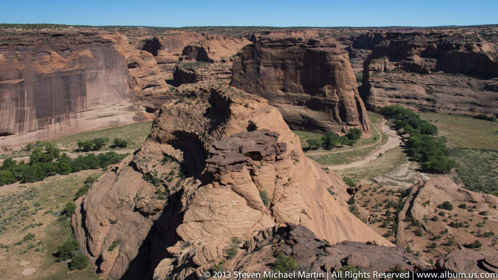 A rounded sandstone outcrop fills the foreground above a broad red-rock canyon with ravines and scattered green vegetation.