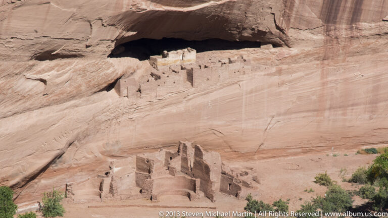 Masonry rooms sit beneath a broad sandstone alcove, with additional structures stepping down the exposed rock slope.