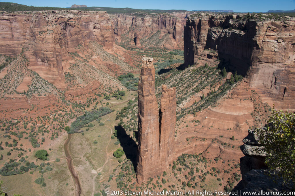 Two slender sandstone spires rise above a green canyon floor, surrounded by expansive red cliffs.