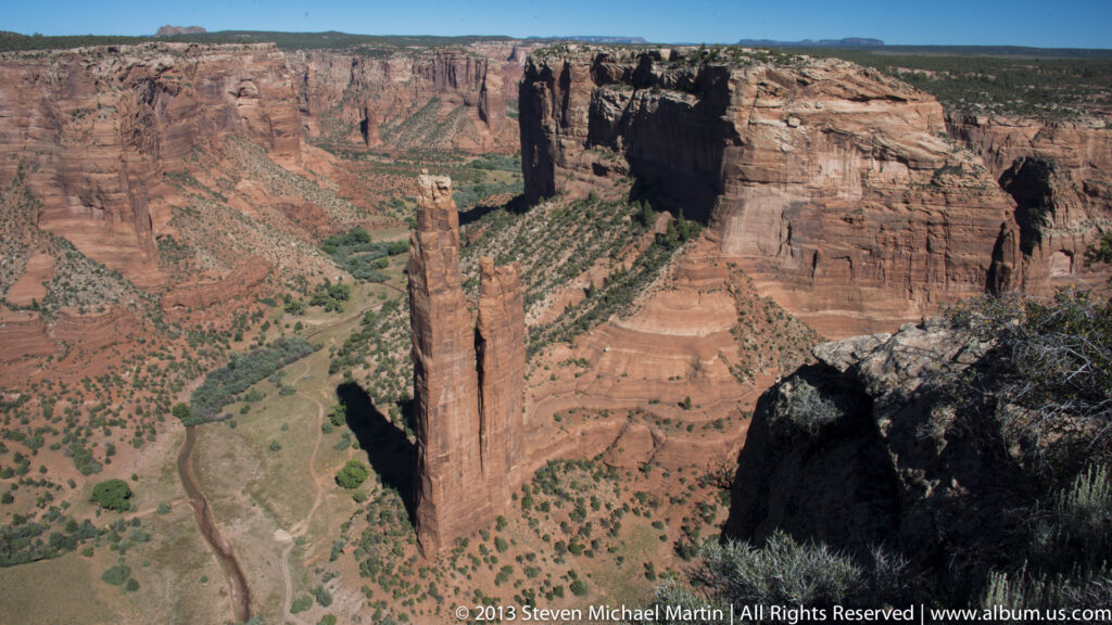 Spider Rock’s twin sandstone towers rise in the distance, partly framed by a dark foreground ledge.