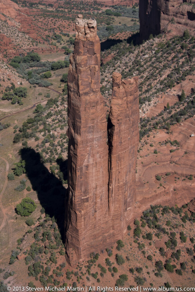 Two sandstone towers with uneven tops cast a long shadow across a scrub-dotted canyon floor.