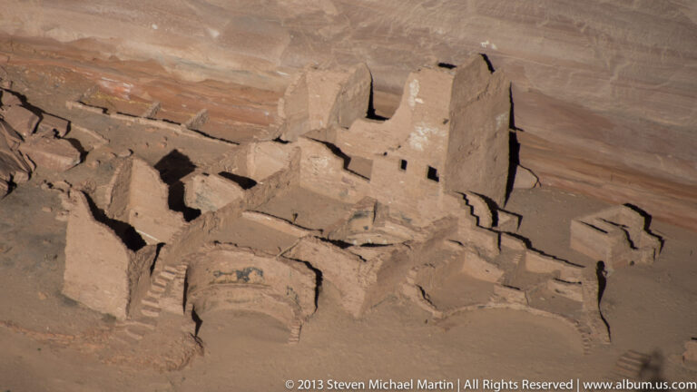 Masonry rooms with rectangular openings cluster on a sloping ledge beneath a massive sandstone cliff.