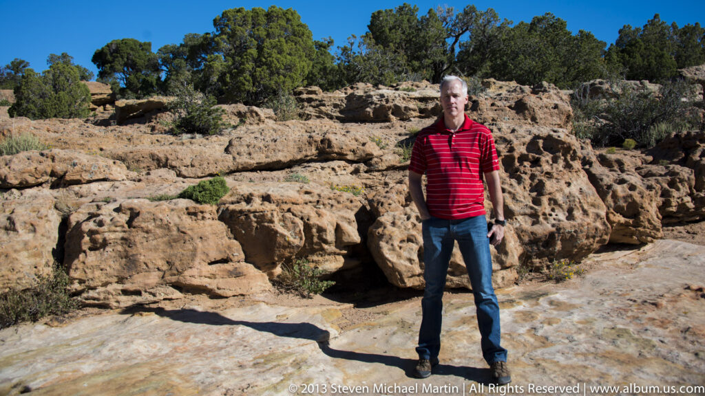 A man in a red striped shirt stands among sandstone blocks, with trees and brush behind him.