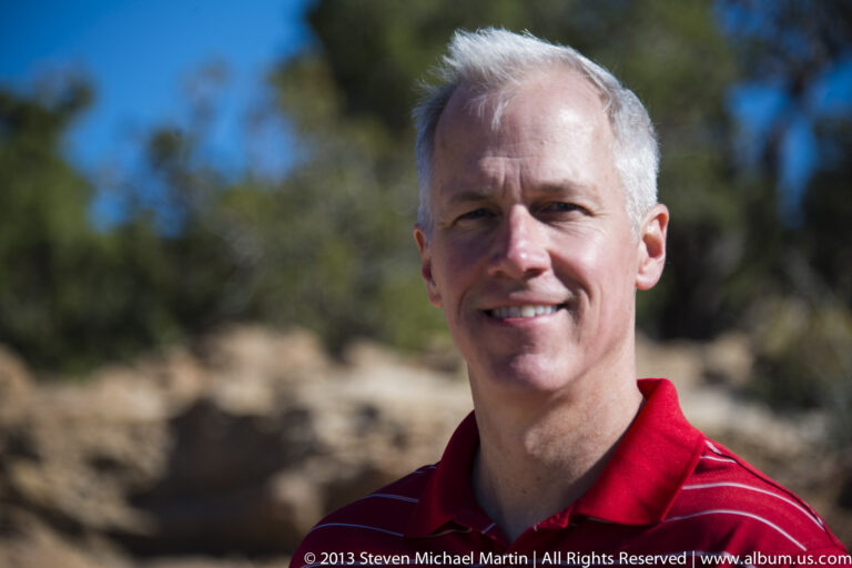 A smiling man with short gray hair wears a red striped polo against blurred sandstone and foliage.