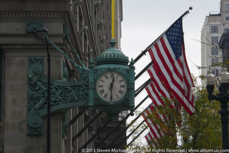 Green ornamental street clock projects from a stone facade beneath several American flags between downtown buildings.