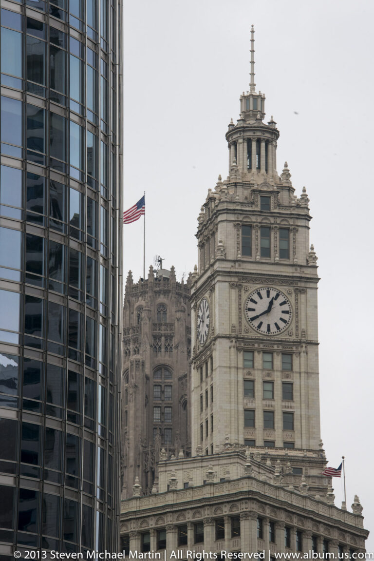 A pale stone clock tower with round clock faces rises beside ornate older facades and a modern glass building.