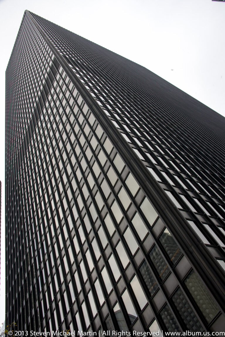 A dark skyscraper facade with a tight window grid narrows toward the sky in a steep upward view.