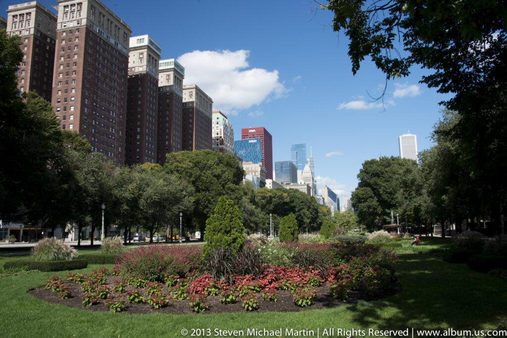 Flower beds and lawn fill a downtown park, with mature trees and Chicago skyscrapers rising behind them.
