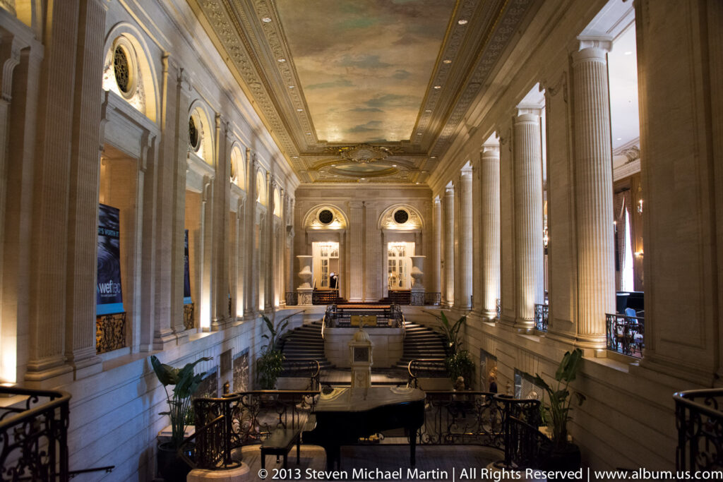 A long public hall with tall columns, dark railings, a central landing, and a painted ceiling recedes toward a doorway.
