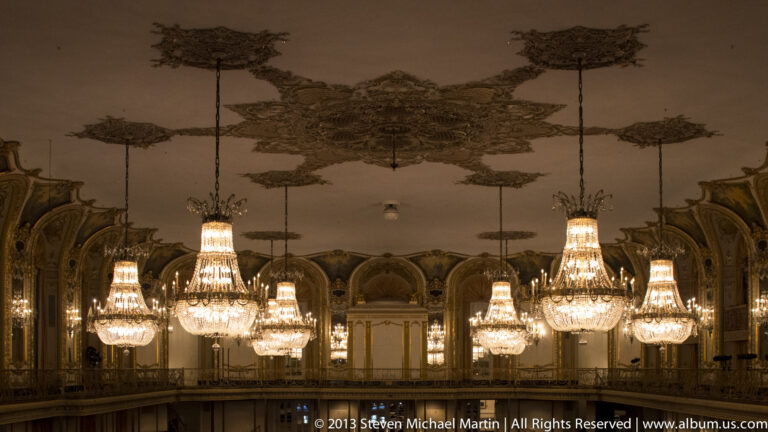 Multiple illuminated chandeliers hang beneath a decorated ceiling with painted panels and repeated arches.
