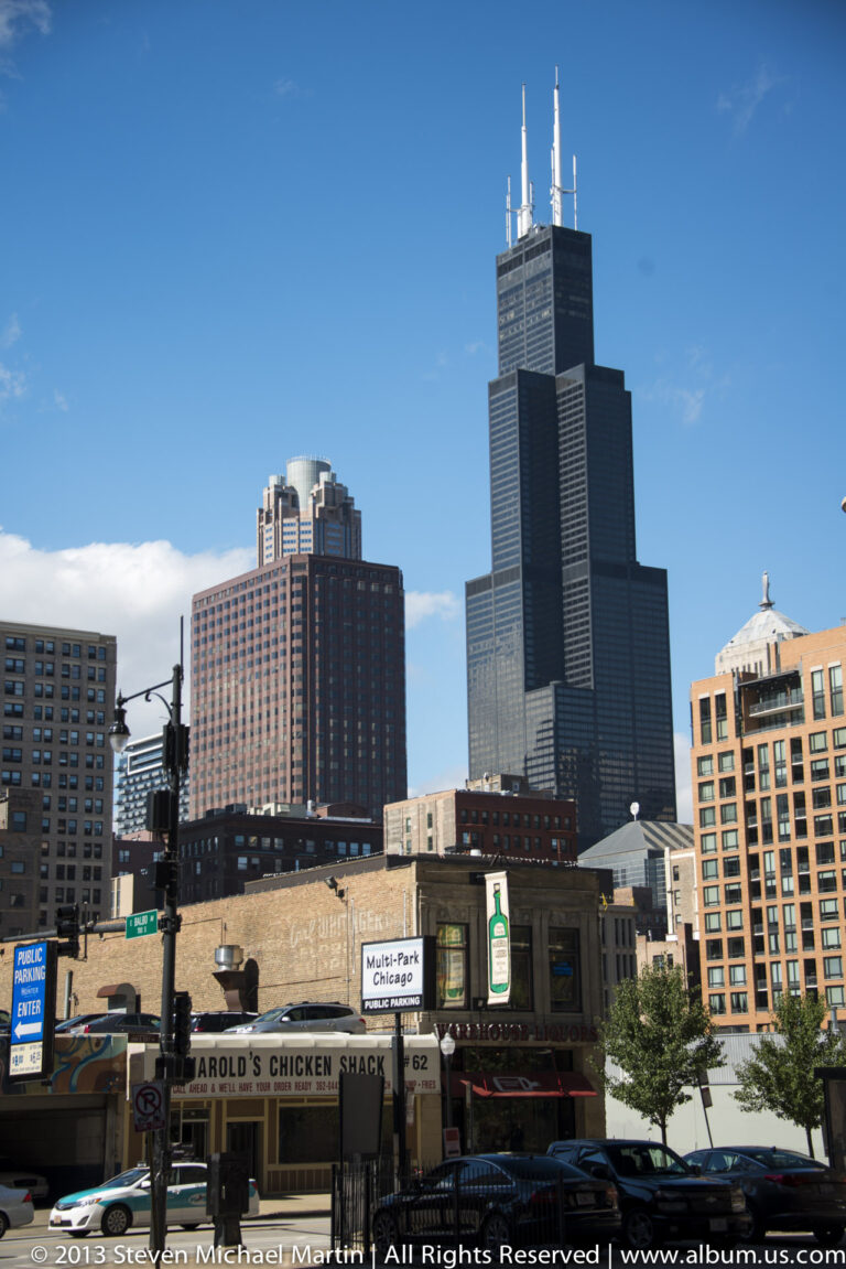 Willis Tower rises behind storefronts and traffic lights on a downtown Chicago street.