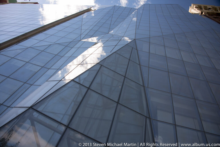 A gridded blue-gray glass facade reflects clouds and buildings, with a bright diagonal streak of light.