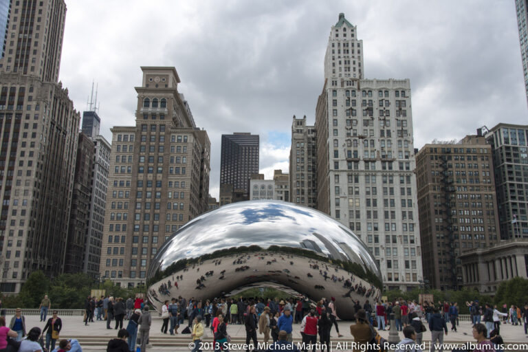 Visitors gather beneath Cloud Gate as it reflects people, clouds, and surrounding buildings across its curved surface.
