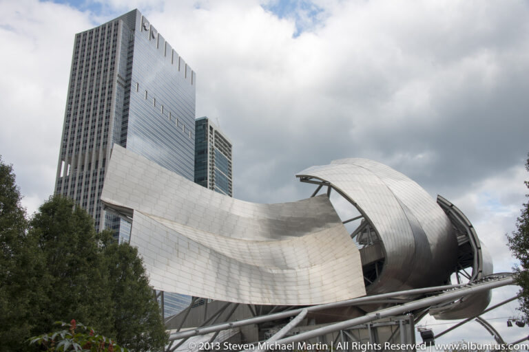 Curving stainless-steel forms and an open canopy define Jay Pritzker Pavilion, with trees and towers behind it.