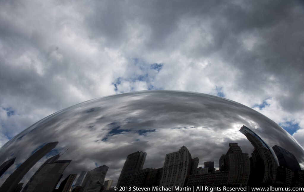Clouds fill the sky above the upper curve of Cloud Gate, which reflects a row of downtown buildings.