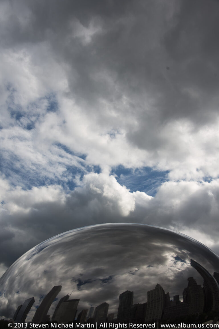 Gray clouds spread above Cloud Gate, whose curved surface reflects dark buildings and a narrow strip of blue sky.