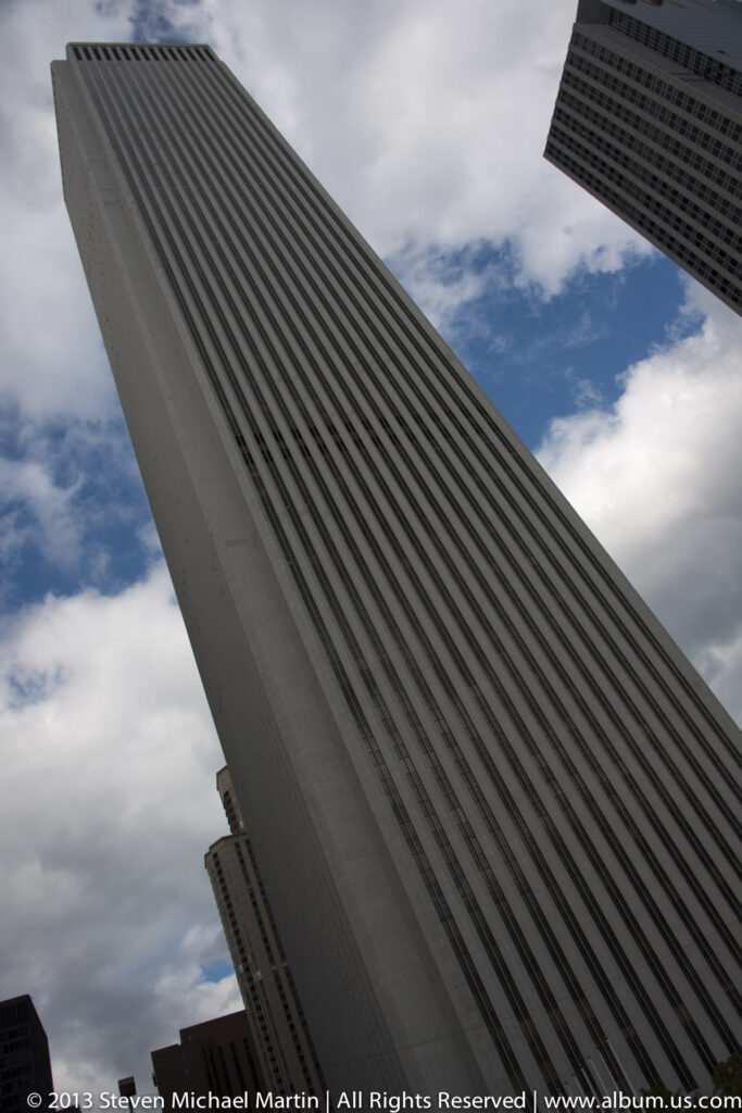 A ribbed dark skyscraper cuts diagonally across the view beneath clouds, with another tower entering at upper right.