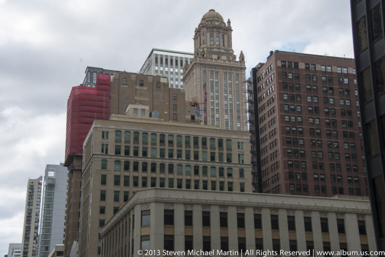 A domed historic tower rises above brick and stone buildings, with red construction covering on a neighboring facade.