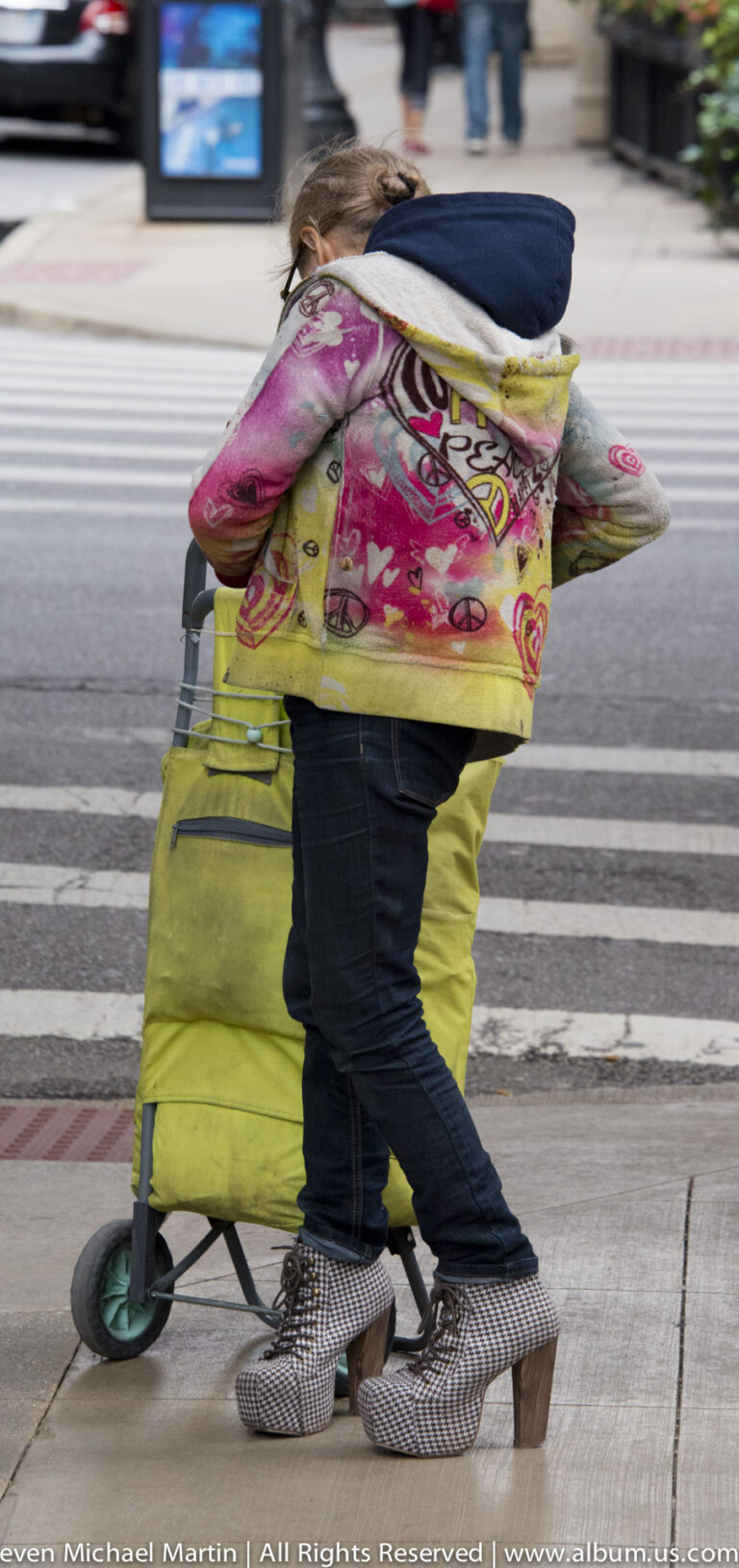 A person in a bright pink-and-yellow jacket moves a yellow wheeled cart across a striped crosswalk.