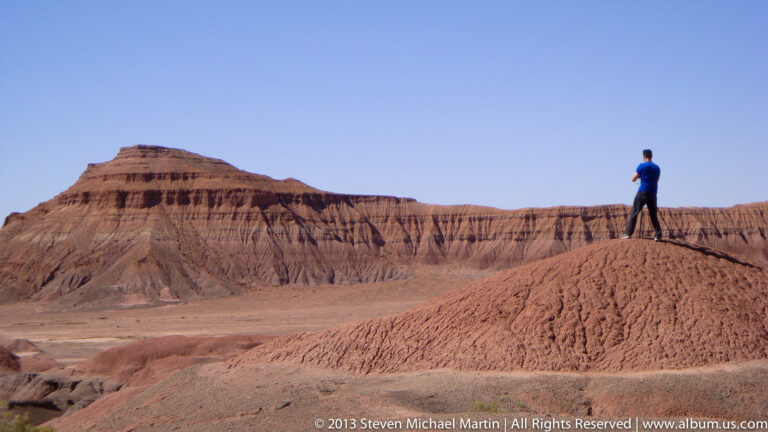 A hiker stands on a narrow red ridge above a basin, with deeply eroded sandstone cliffs extending across the background.