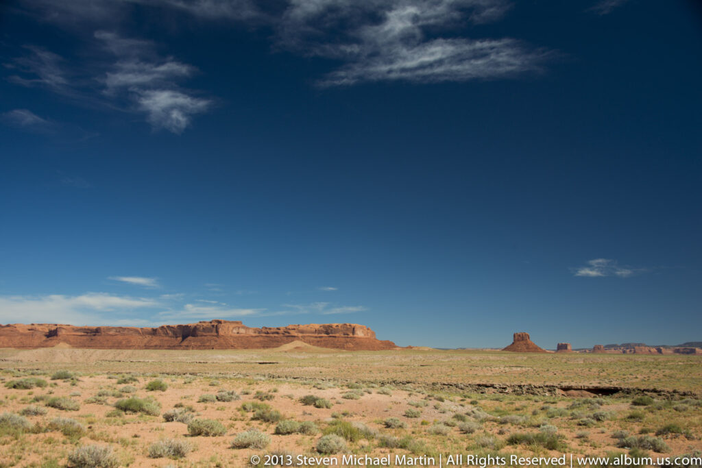 A wide desert plain with scattered shrubs stretches toward distant mesas and isolated buttes beneath a deep blue sky.