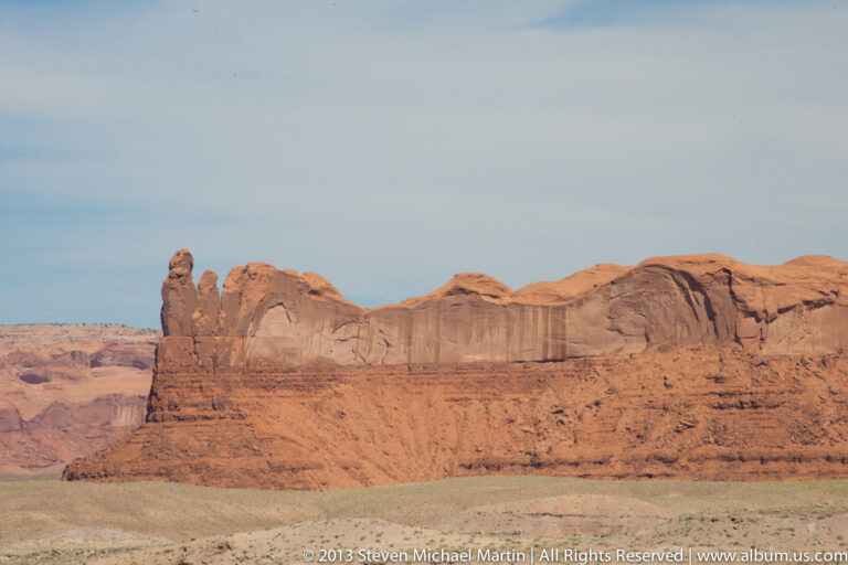 A narrow sandstone spire rises before a long red escarpment, with rounded domes and rock shelves extending to the right.