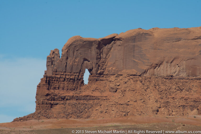 A dark opening cuts through a projecting sandstone buttress beneath a broad red rock wall and blue sky.