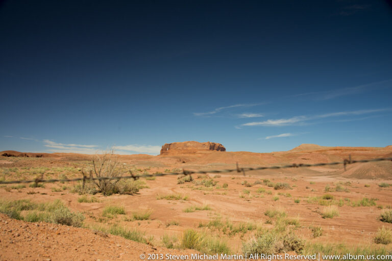 A distant red mesa rises above a sandy plain dotted with shrubs, with a thin dark line crossing the foreground.